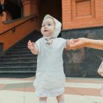 Adorable baby in a white christening outfit being held outdoors on a warm day.