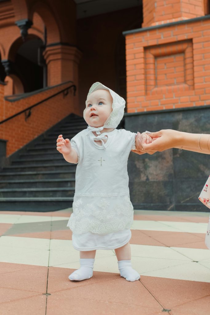 Adorable baby in a white christening outfit being held outdoors on a warm day.